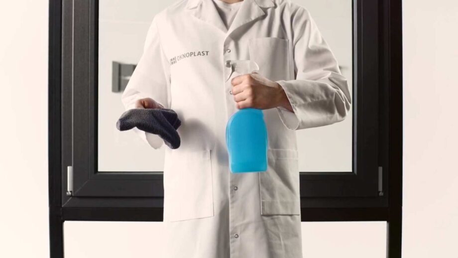 Un expert OKNOPLAST en blouse blanche tenant dans la main un produit d'entretien fenêtre et un chiffon doux, avec une fenêtre dans le fond