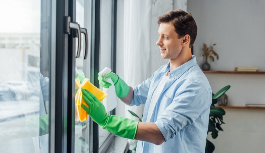 Un homme en chemise bleue et gants verts qui nettoie les vitres d'une fenêtre avec un chiffon doux jaune