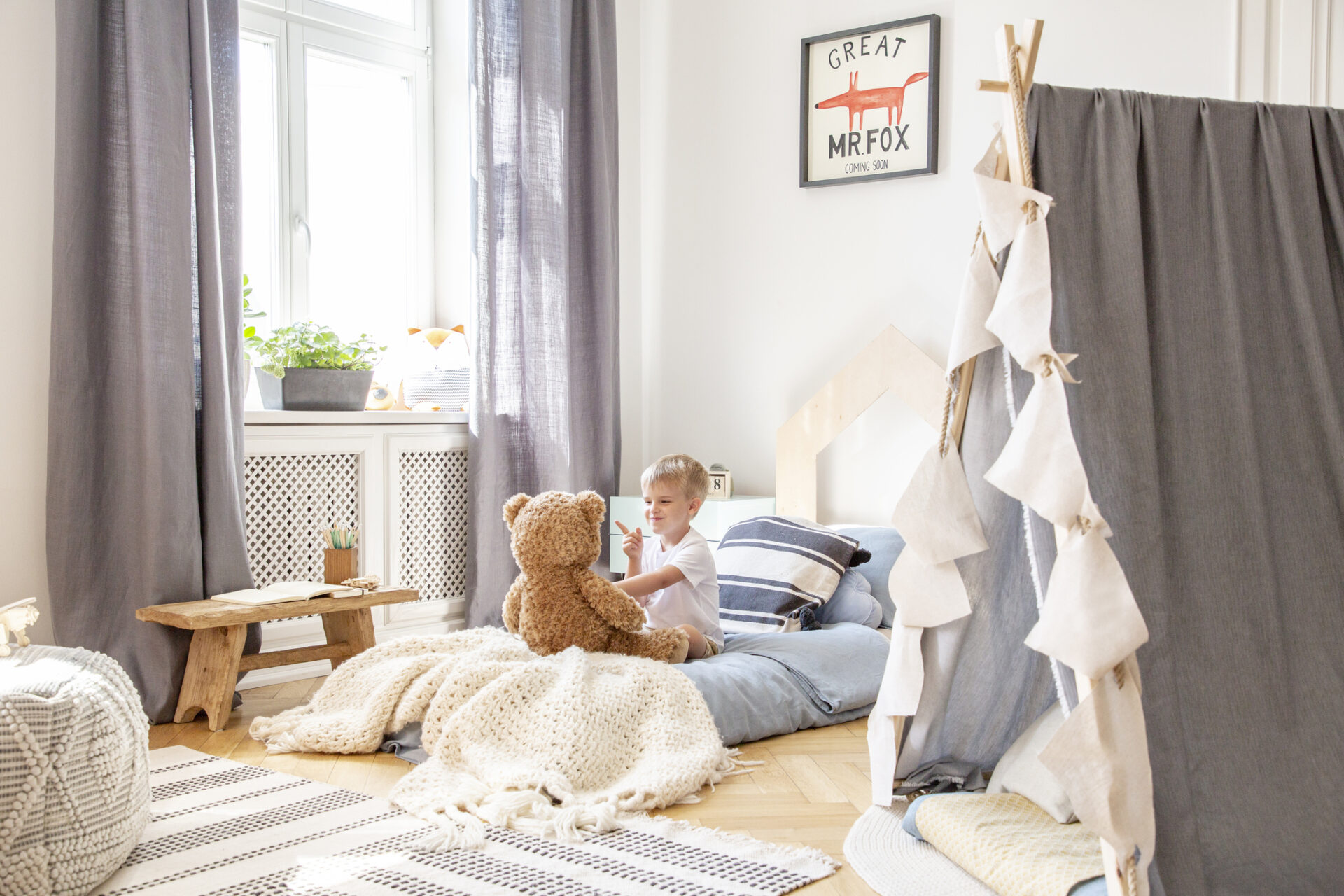 Un petit garçon assis sur son lit joue avec son nounours dans sa chambre, avec une fenêtre blanche, rideaux gris et une tente en lin gris