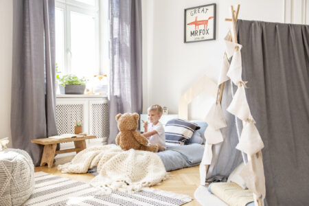 Un petit garçon assis sur son lit joue avec son nounours dans sa chambre, avec une fenêtre blanche, rideaux gris et une tente en lin gris